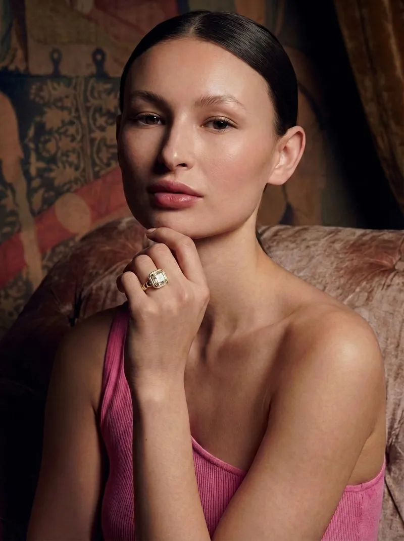 FormaNova AI's fine jewelry product photo featuring a model wearing a prominent, square-set bezel ring in yellow gold. She rests her chin on her hand, wearing a pink ribbed top against a textured, antique armchair and tapestry backdrop.